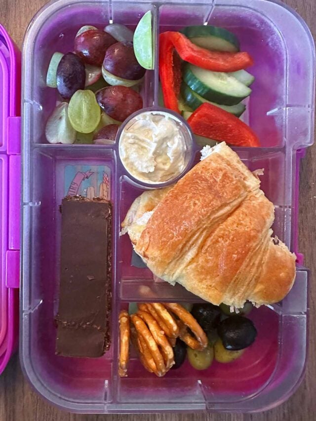 A compartment lunchbox containing half a croissant, fruit, veg and a chocolate bar.