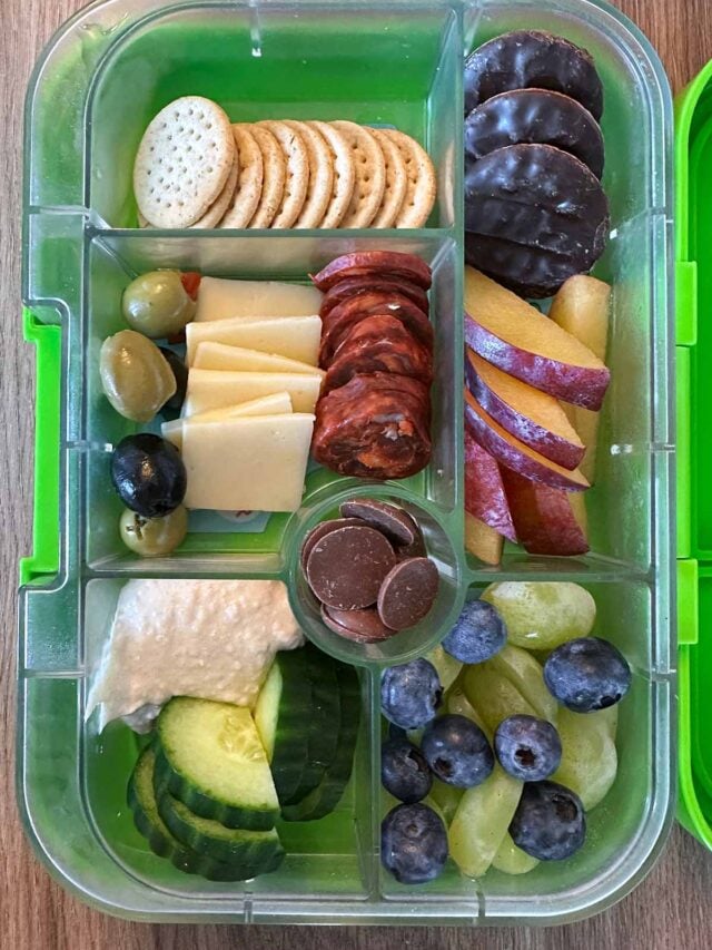 A compartment lunchbox containing crackers, cheese, slices chorizo, berries, peach slices and mini jaffa cakes.