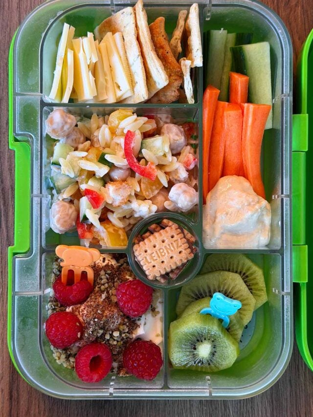 A compartment lunchbox containing pasta salad, fruit topped yogurt, cheese, crackers, crudités and kiwi.