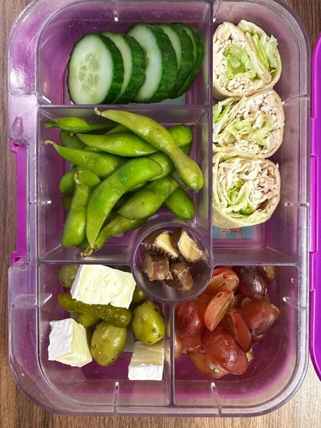 A compartment lunchbox containing a wrap, edamame, cucumber, grapes, cheese, pickles and chocolate.