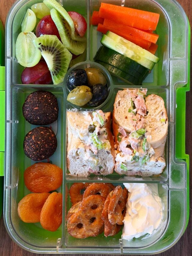 A compartment lunchbox containing a bagel, fruit, veg, pretzels, cream cheese and energy balls.