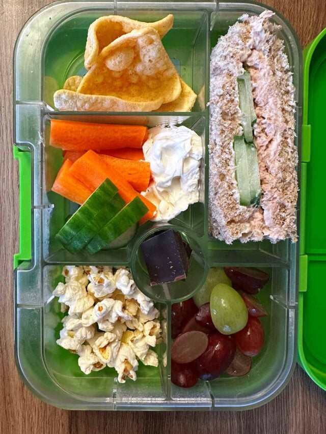 A compartment lunchbox containing a sandwich, crisps, grapes, popcorn, crudités and chocolate.