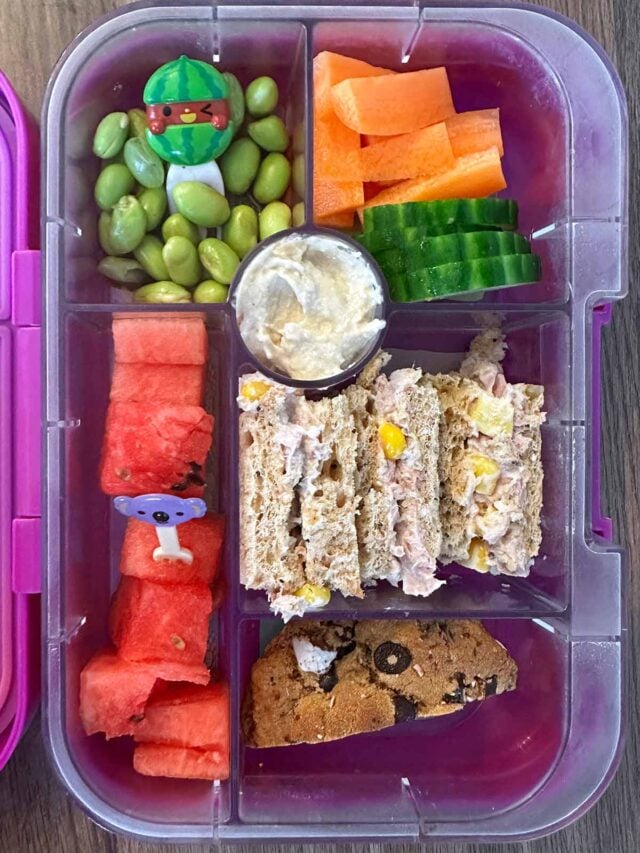 A compartment lunchbox containing a sandwich, veg, edamame, watermelon and a muffin.
