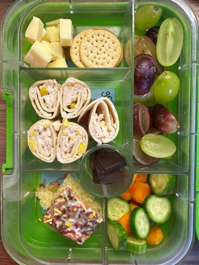 A compartment lunchbox containing wraps, fruit, veg, cheese, crackers and school cake.