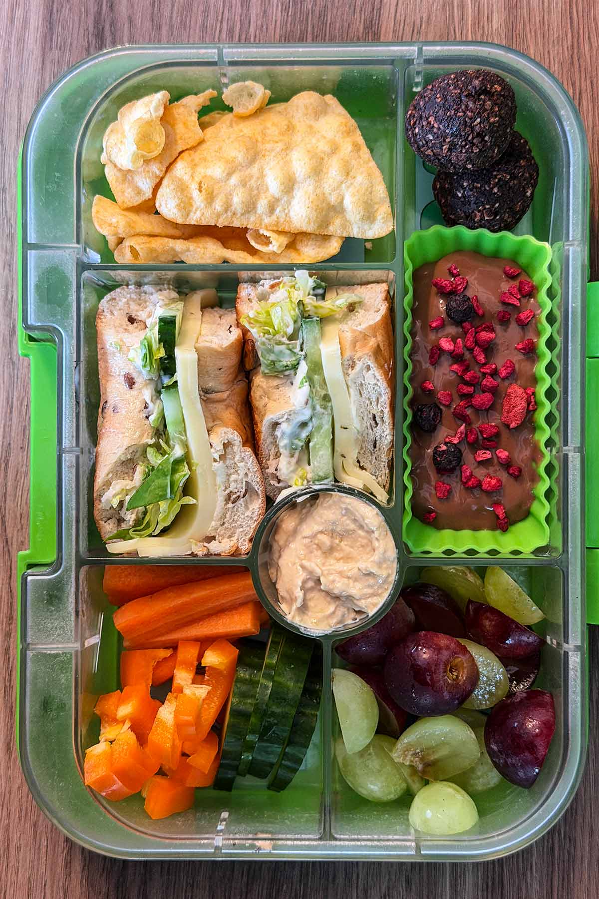 A compartment lunchbox containing a cheese salad bagel, crudités, hummus, grapes, crisps and chocolate pudding.