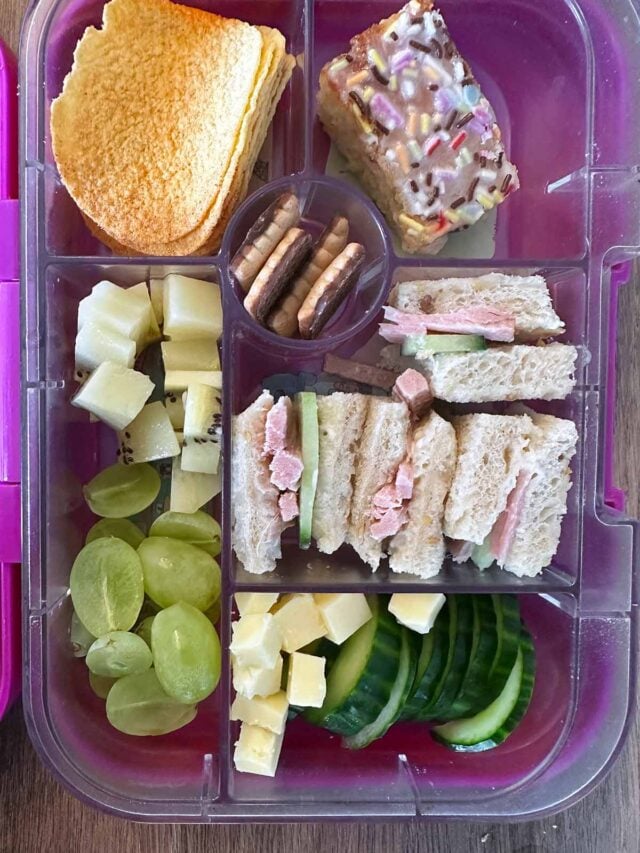 A compartment lunchbox containing a sandwich, cucumber, cheese, grapes, kiwi, crisps and school cake.