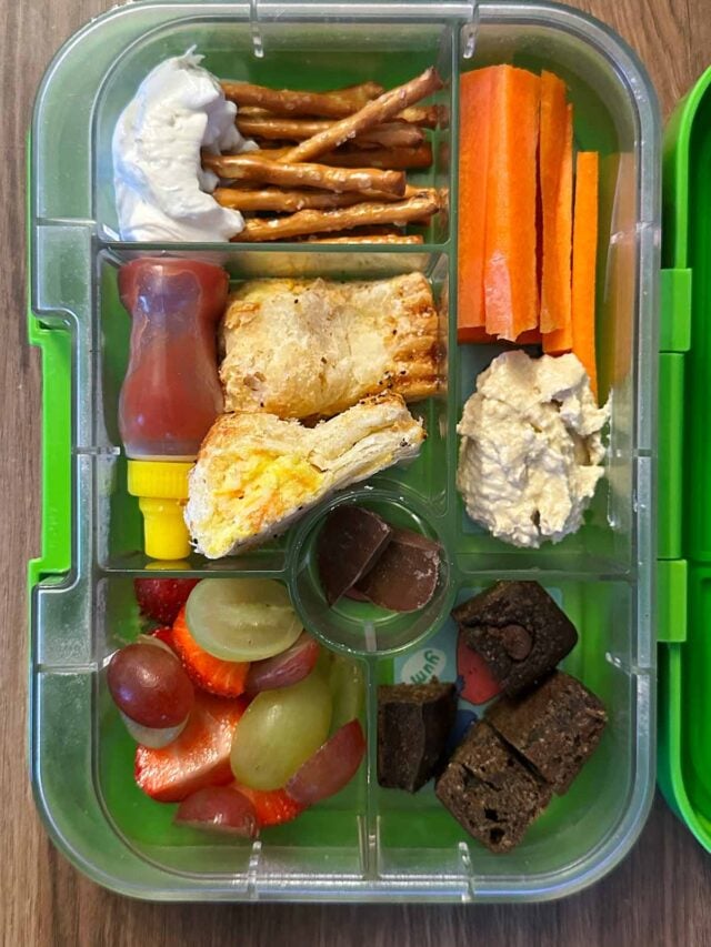 A compartment lunchbox containing cheese roll, pretzels, dip, carrot, fruit and a brownie.