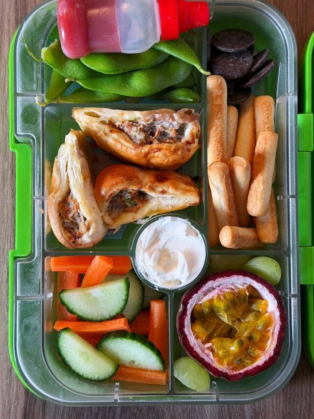 A compartment lunchbox containing sausage rolls, edamame, carrot, cucumber, passionfruit and chocolate buttons.