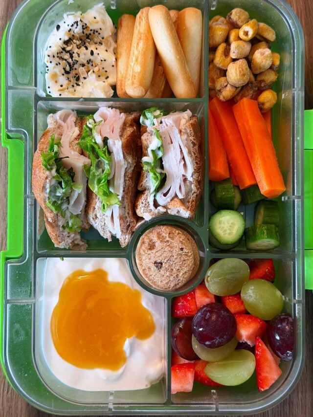 A compartment lunchbox containing a bagel, hummus, breadsticks, corn, fruit, veg and yogurt.