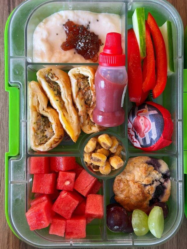A compartment lunchbox containing sausage rolls, rice pudding, fruit, veg, corn, cheese and blueberry muffin.