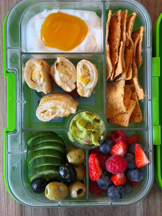 A compartment lunchbox containing cheese roll, crisps, guacamole, fruit, veg and yogurt.