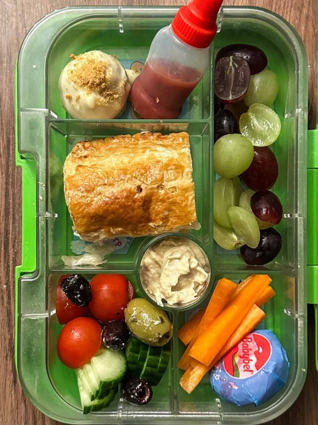 A compartment lunchbox containing sausage roll, grapes, cheese and salad veg.
