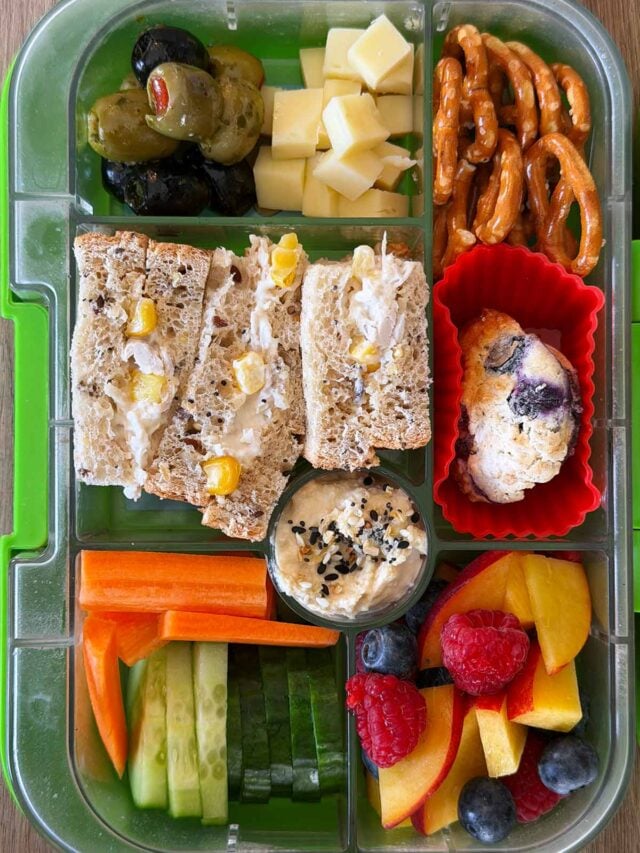 A compartment lunchbox containing a chicken and sweetcorn sandwich, fruit, veg, cheese, pretzels and a muffin.