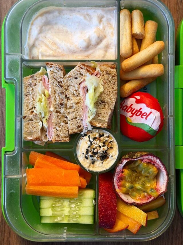 A compartment lunchbox containing a ham sandwich, veg, breadsticks, hummus, cheese, passionfruit and yogurt.