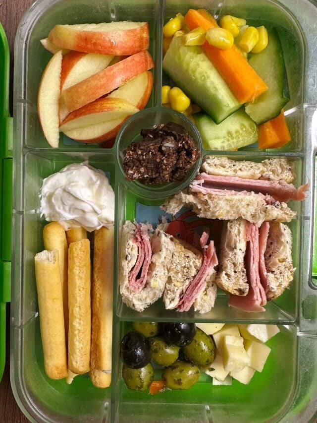 A compartment lunchbox containing a ham sandwich, fruit, veg, olives, cheese, breadsticks and cream cheese.