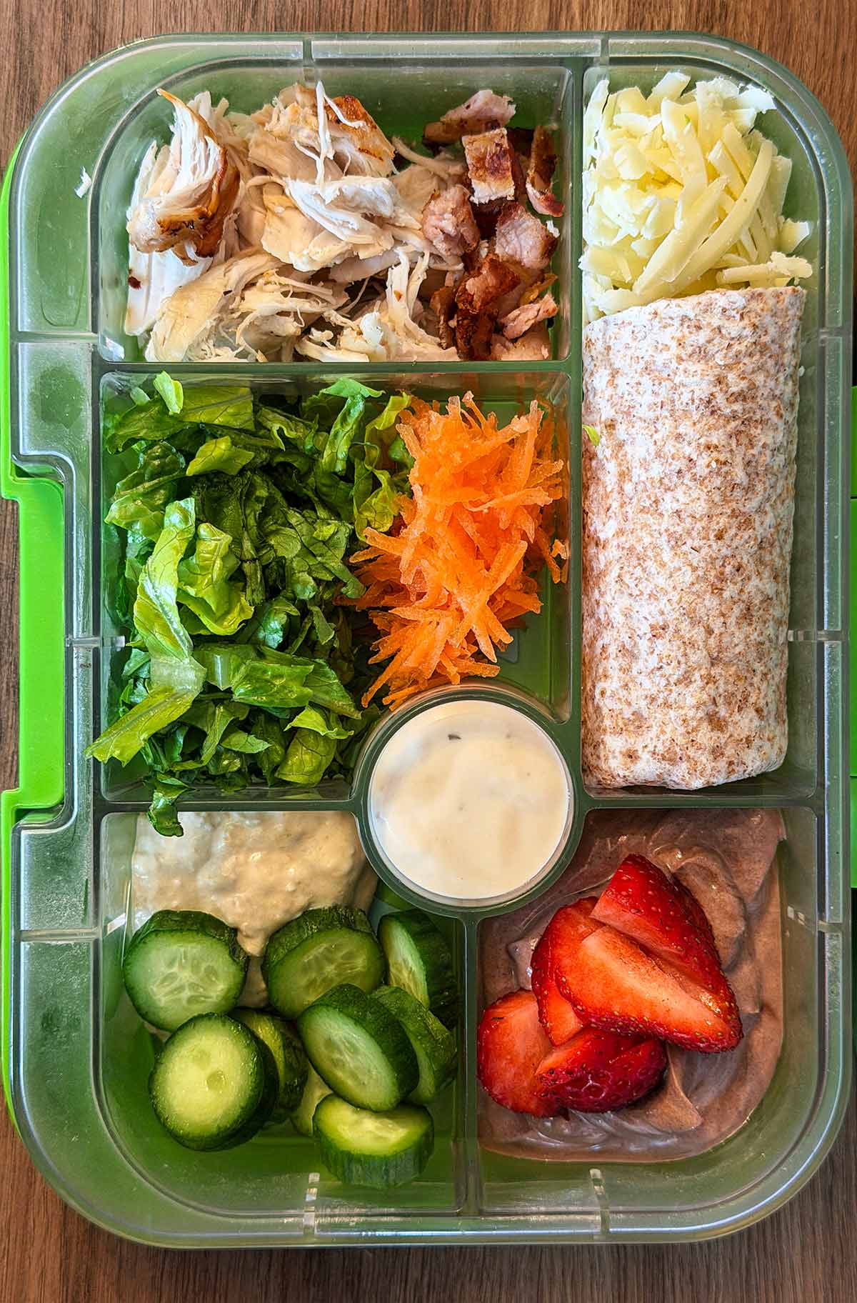 A compartment lunchbox containing a wrap, lettuce, grated carrot, shredded chicken, grated cheese, Caesar sauce, cucumber slices, hummus and fruit topped chocolate pudding.