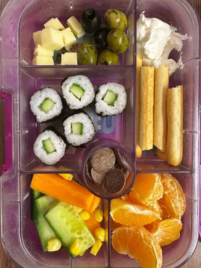 A compartment lunchbox containing sushi, fruit, veg, olives, cheese, breadsticks and chocolate buttons.