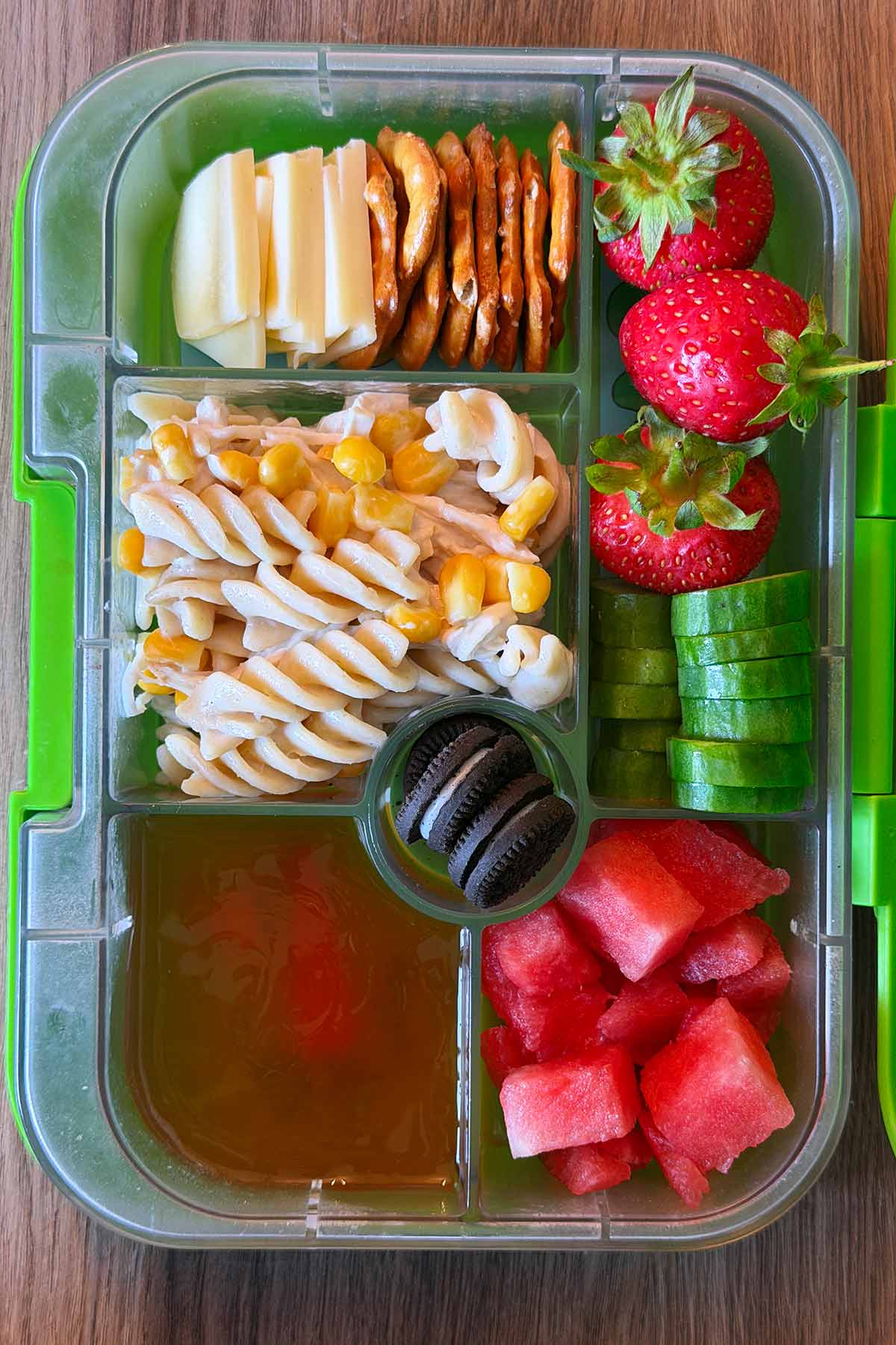 A compartment lunchbox containing pasta salad, cheese and crackers, fruit, veg, jelly and mini oreos.