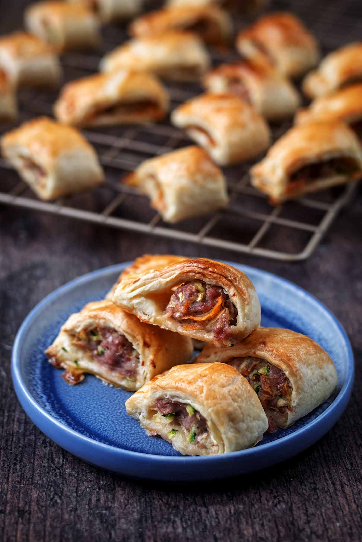 A plate of sausage rolls in front of a cooling rack with more sauasge rolls.