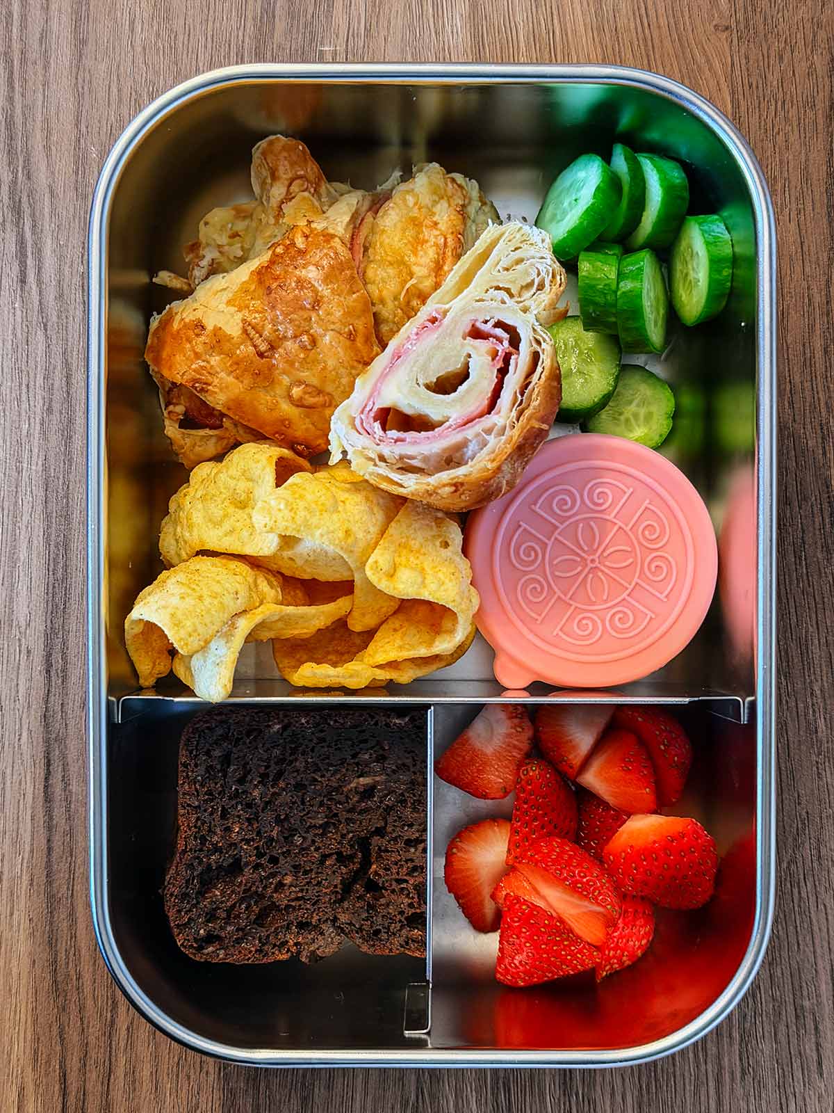 A lunchbos with mini croissants, vegetables, berries and a small slice of chocolate cake.