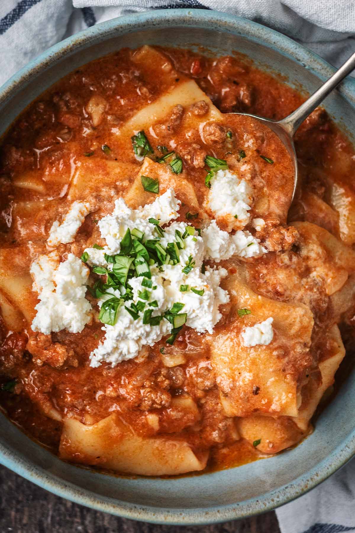 A bowl of lasagna soup topped with crumbled ricotta cheese and chopped herbs.